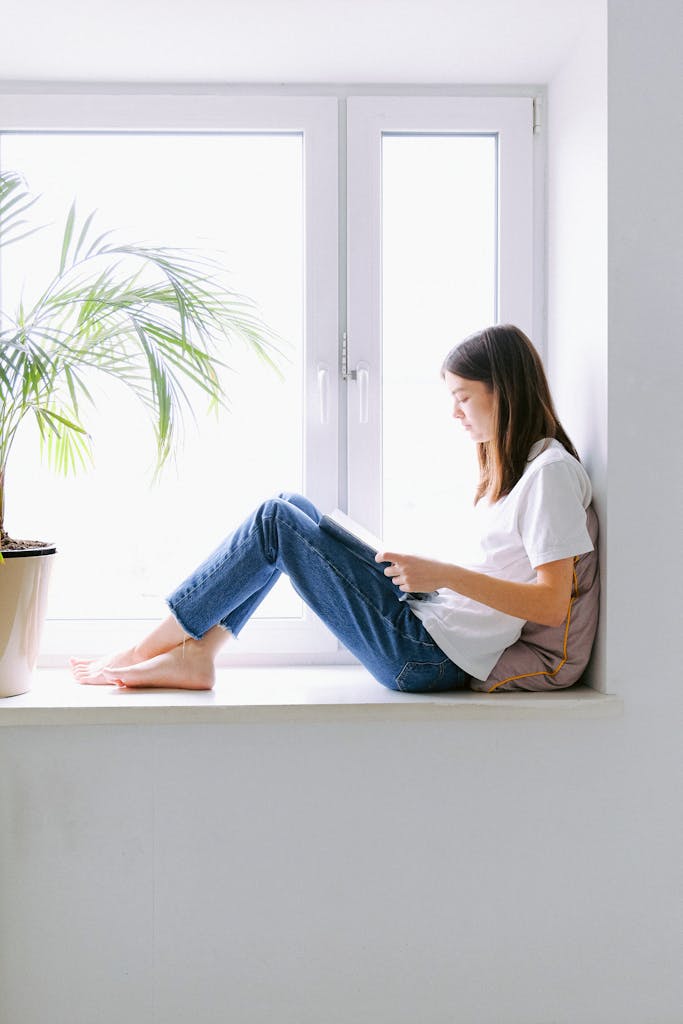 Teen girl enjoying a book while sitting by a bright window indoors.